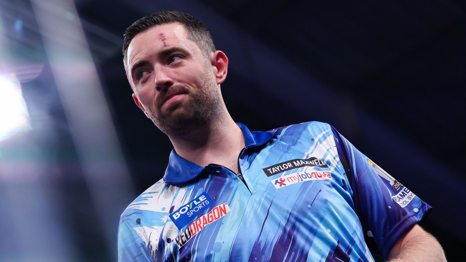 A bearded darts player in a blue patterned shirt looks upward under bright arena lights.