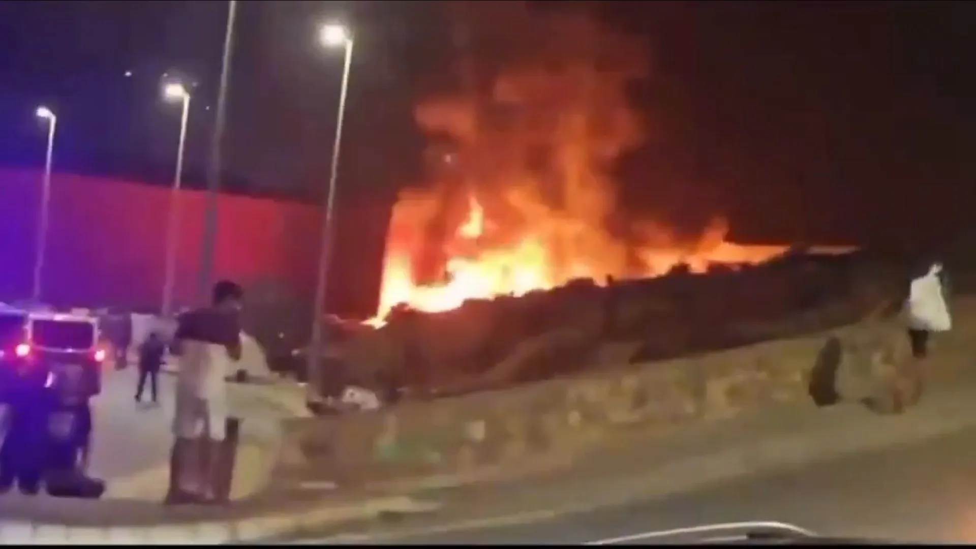 Nighttime fire engulfs a large structure as emergency vehicles and people stand nearby.