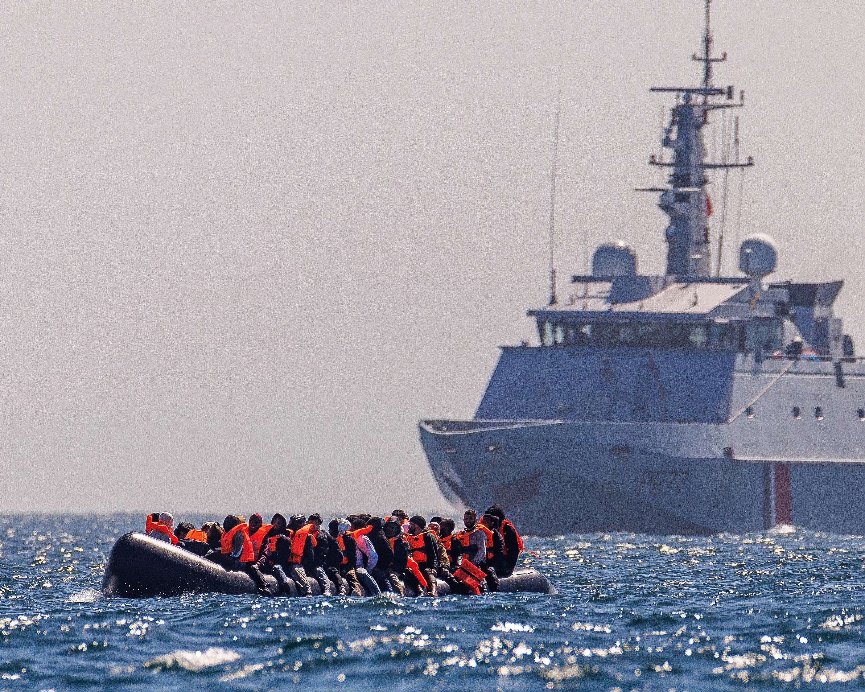 People in orange life jackets sit in an inflatable boat near a large patrol vessel at sea.