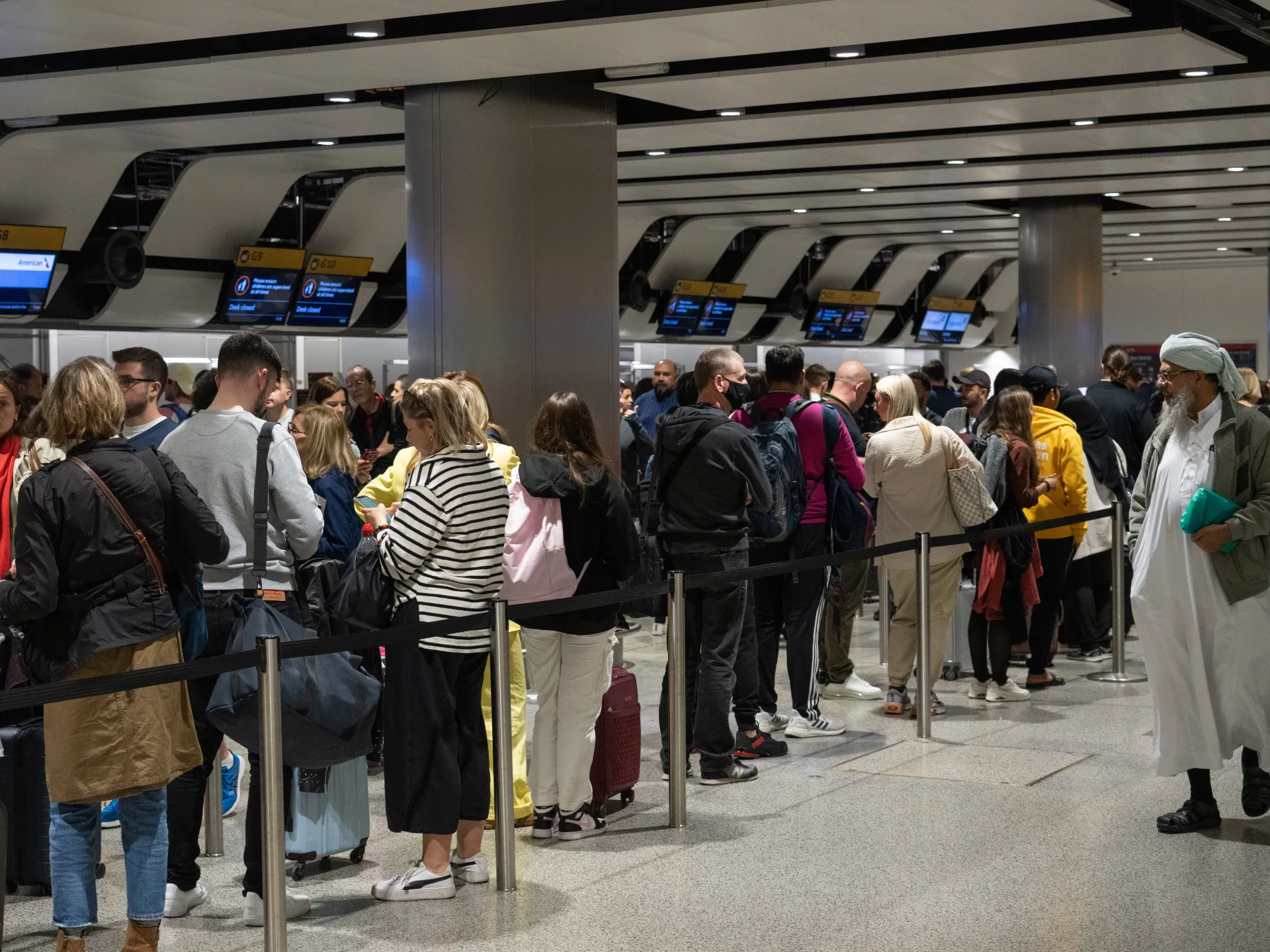 Passengers queue inside a crowded airport terminal beneath gate screens and ceiling panels.