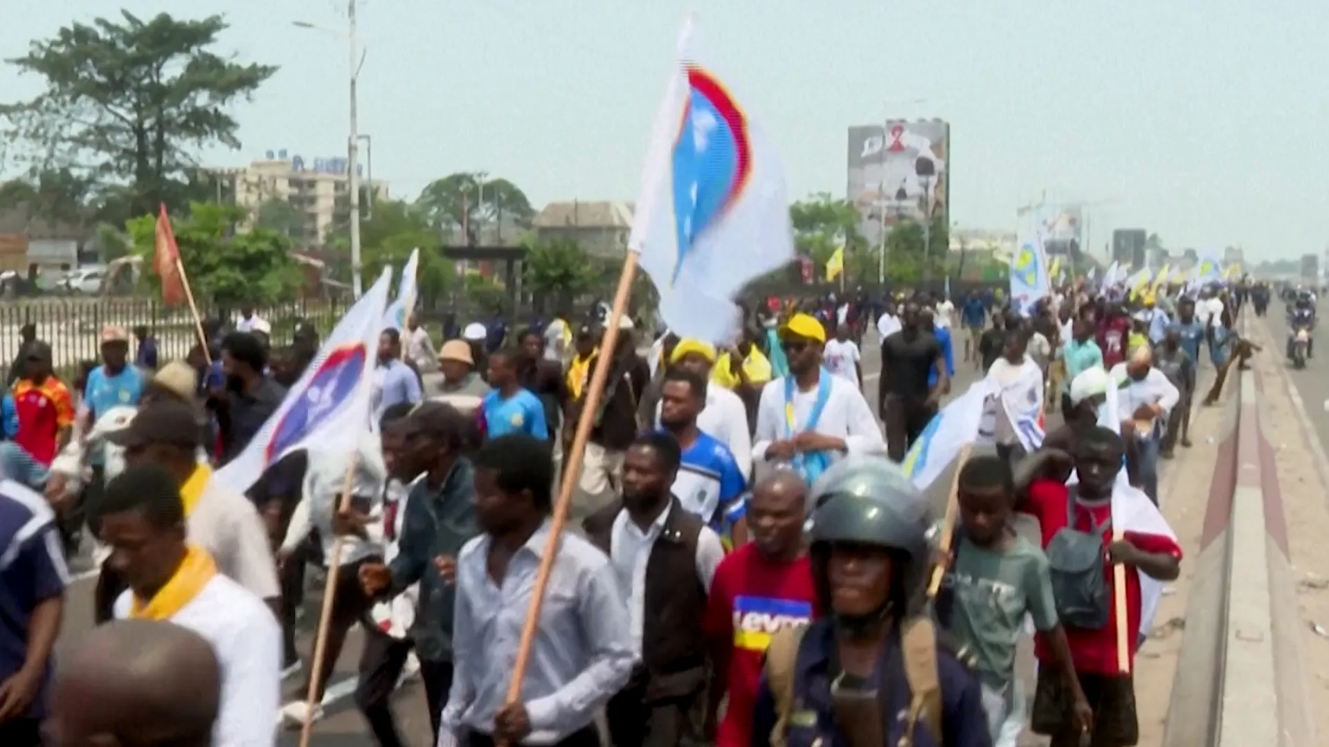 Large crowd marching along a roadway while carrying white, yellow, and multicolored flags