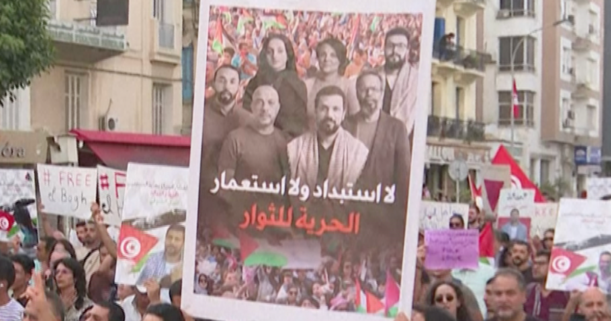 Protesters hold signs and Tunisian flags, including a large Arabic-language poster, in a crowded demonstration.