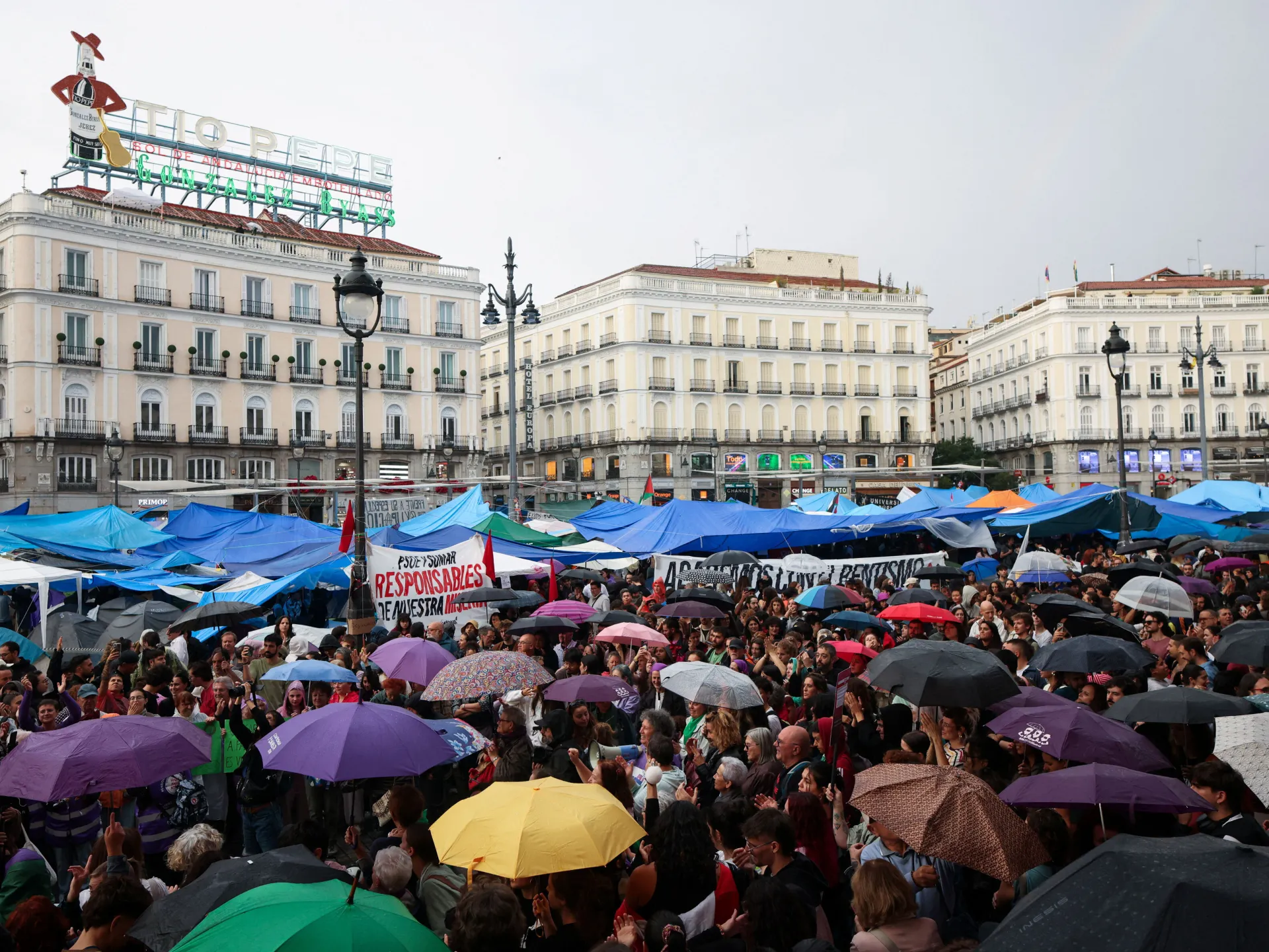 Crowd beneath blue tent canopies and colorful umbrellas in a city square, with historic buildings behind.