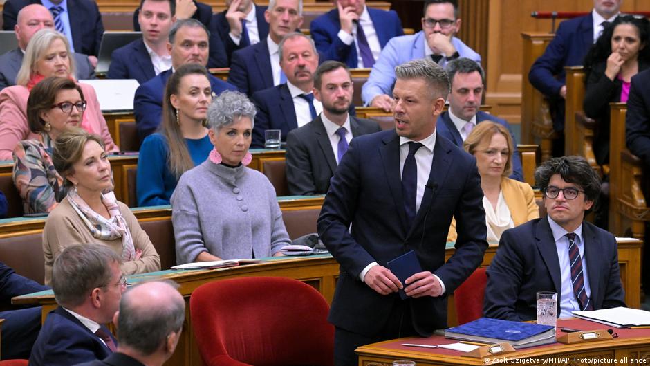 A suited man speaks in a parliamentary chamber as lawmakers sit around him.