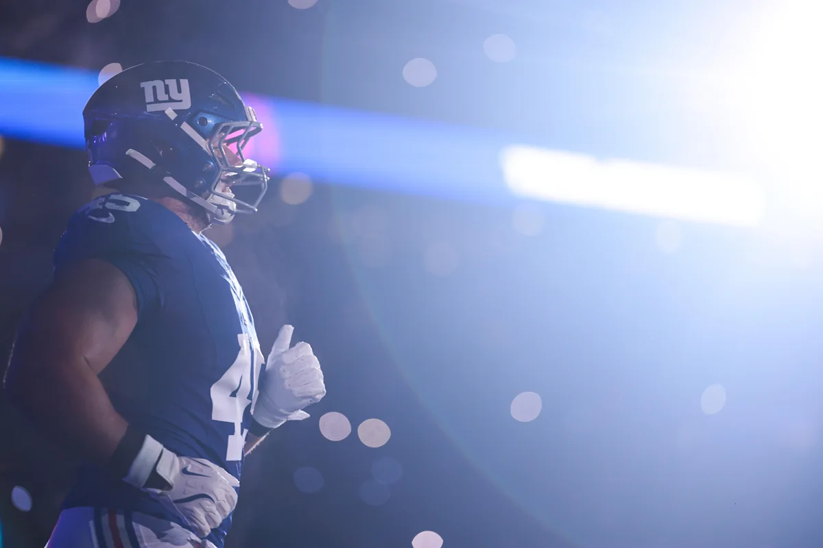 A Giants football player in uniform runs through blue stadium lights, wearing a helmet and gloves.