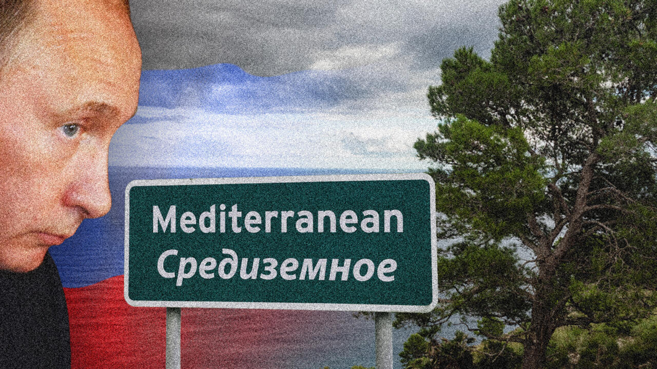 Close-up of a man beside a Mediterranean sign, with a Russian flag and pine trees in the background.