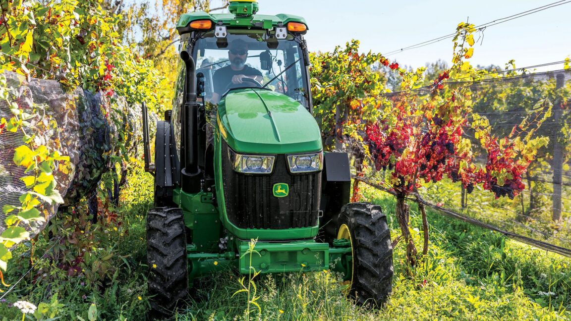 Green John Deere tractor parked between rows of grapevines on a sunny farm