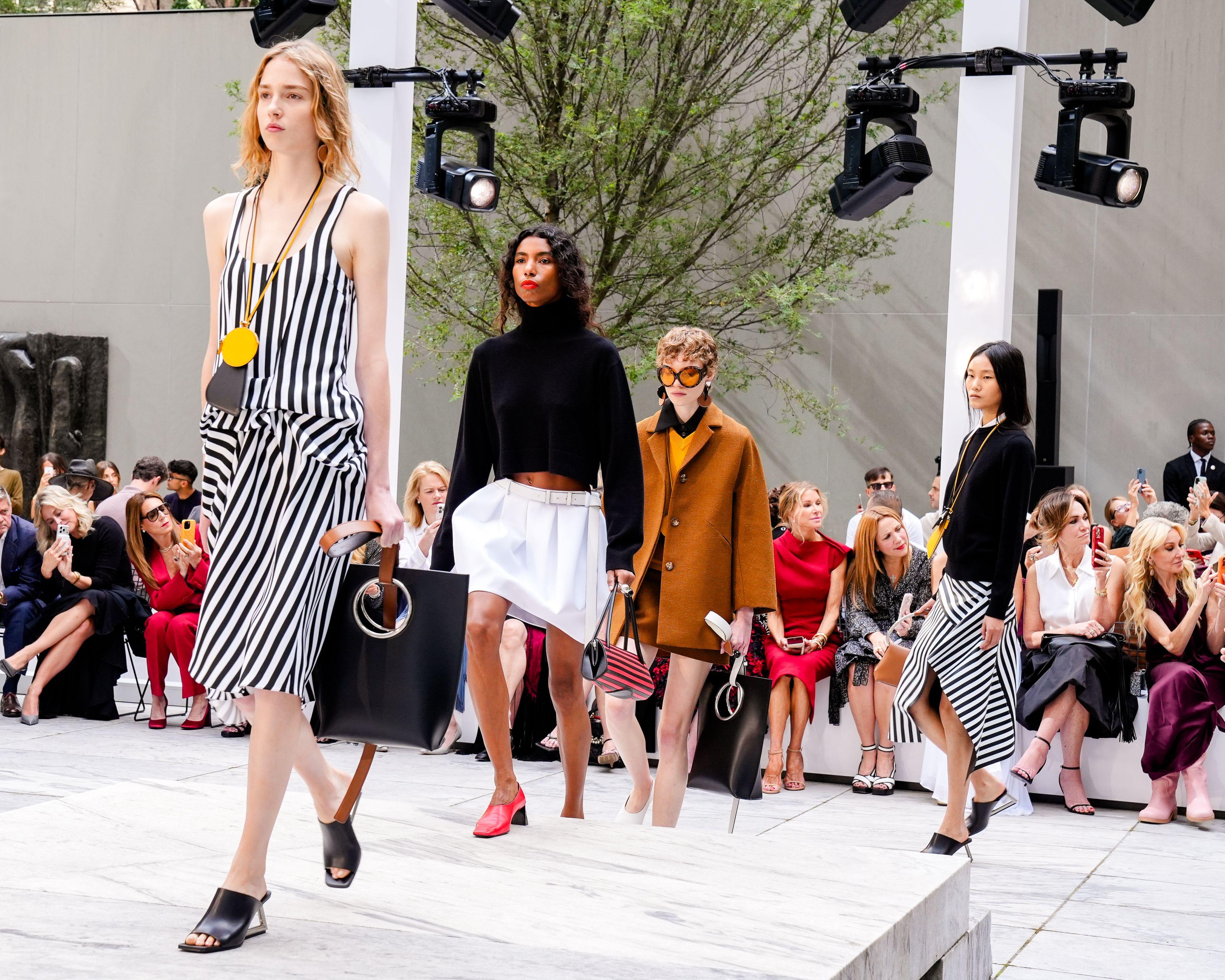 Models walk a runway in striped outfits and colorful tailoring before a seated audience.