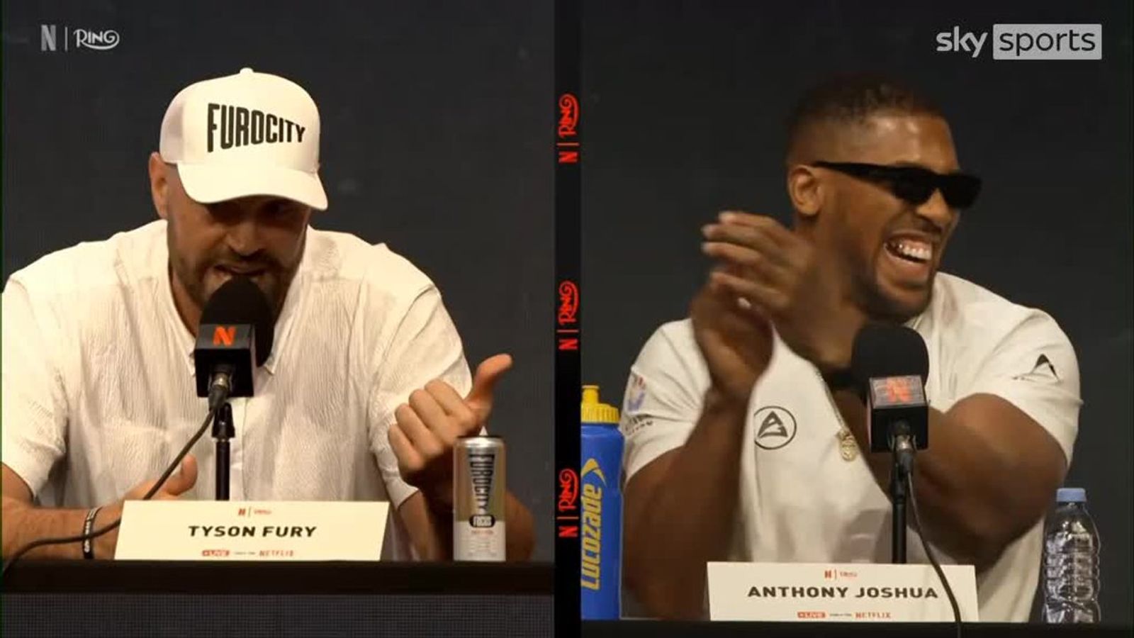 Tyson Fury speaks into a microphone as Anthony Joshua laughs and claps beside him at a press table.
