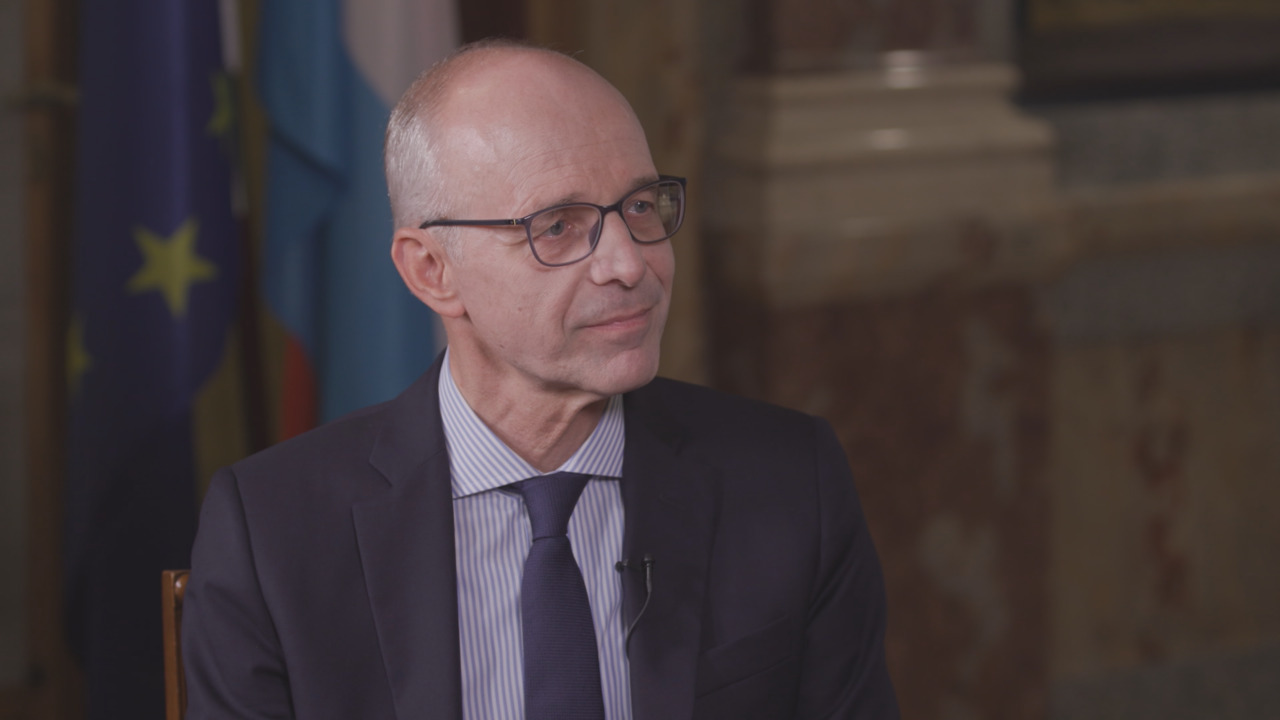 Bald man wearing glasses and a dark suit sits before European flags and a stone wall.