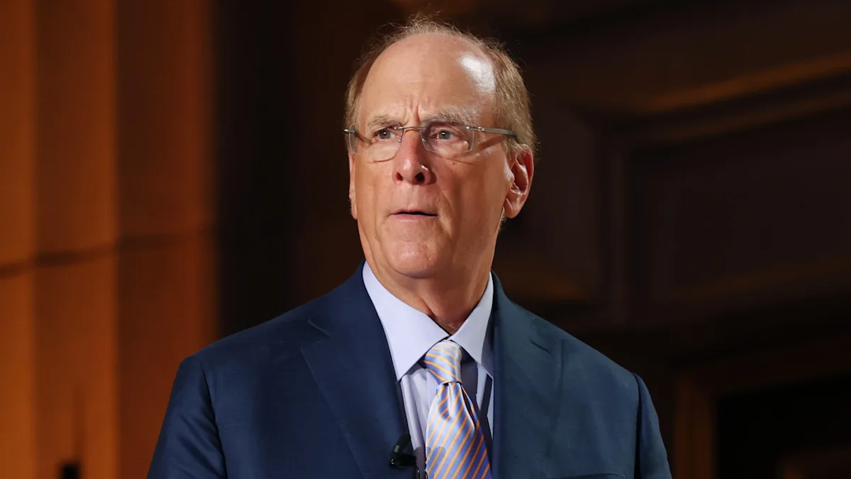 Older man in glasses and dark suit speaking indoors under warm lighting.