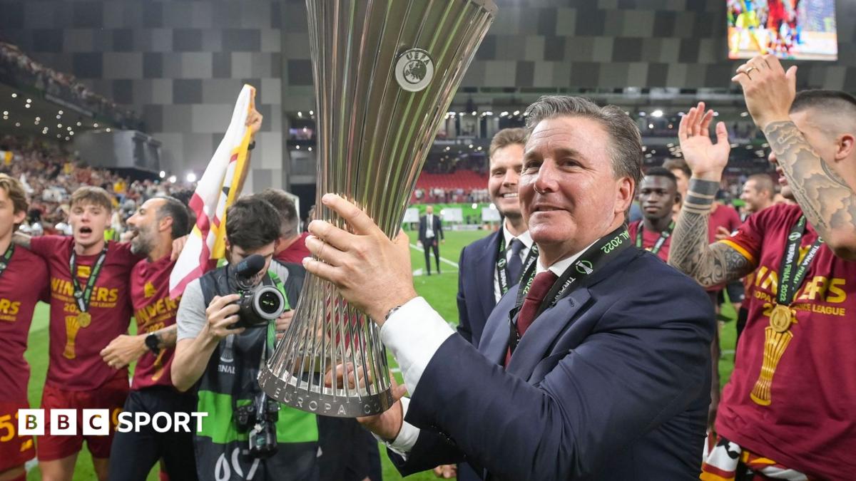 Man in suit holds a large trophy as football players celebrate around him