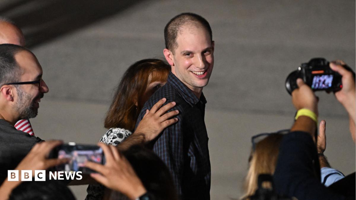 A smiling man in a dark checked shirt is surrounded by people holding cameras and phones.