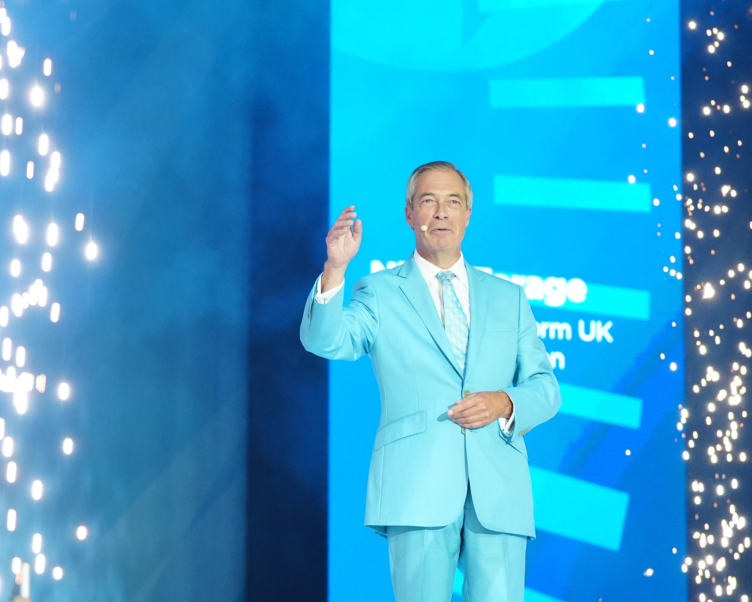 Man in pale blue suit speaking onstage with one hand raised amid bright blue lights