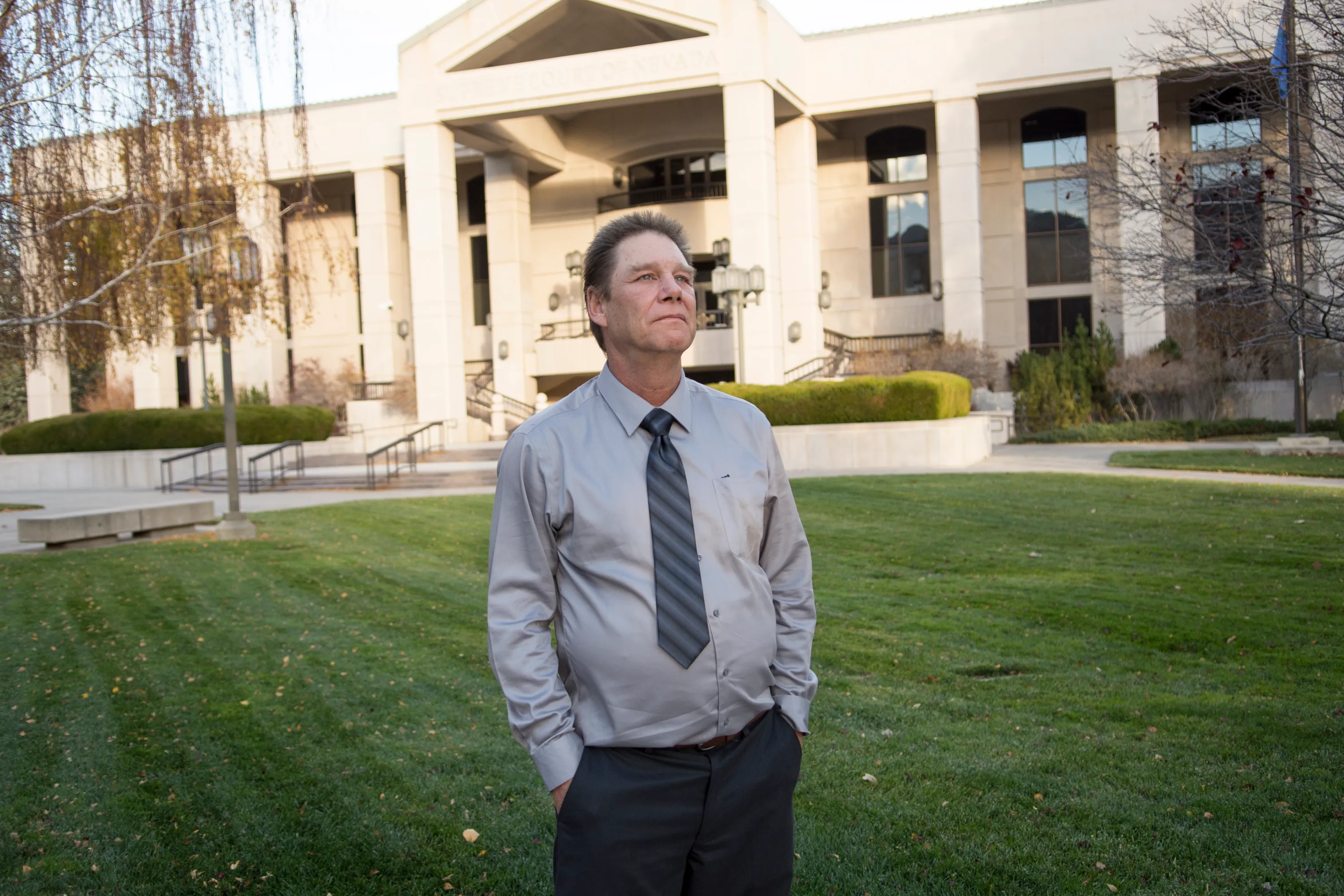 Man in dress shirt and tie standing on a lawn outside a large institutional building.