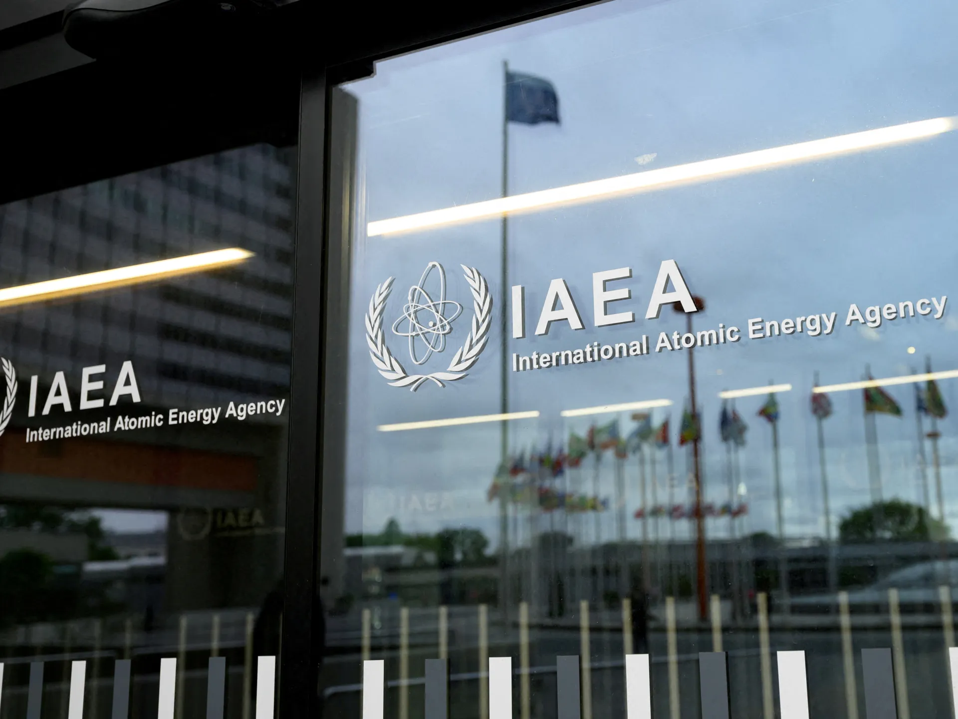 IAEA logo displayed on a glass entrance reflecting flags, buildings, and interior lighting.