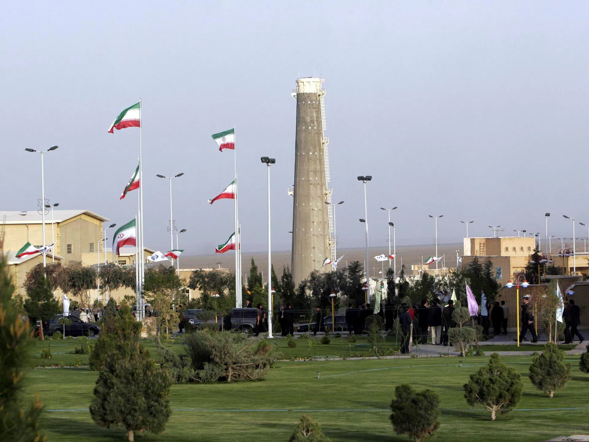 Iranian flags fly around a tall industrial tower and landscaped grounds under a hazy sky.