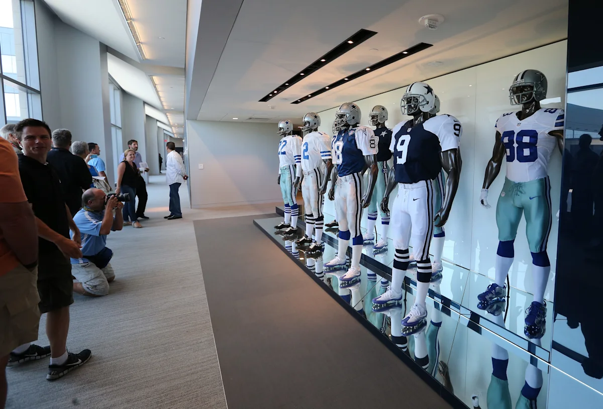 Mannequins display Dallas Cowboys football uniforms in a bright hallway as visitors observe.
