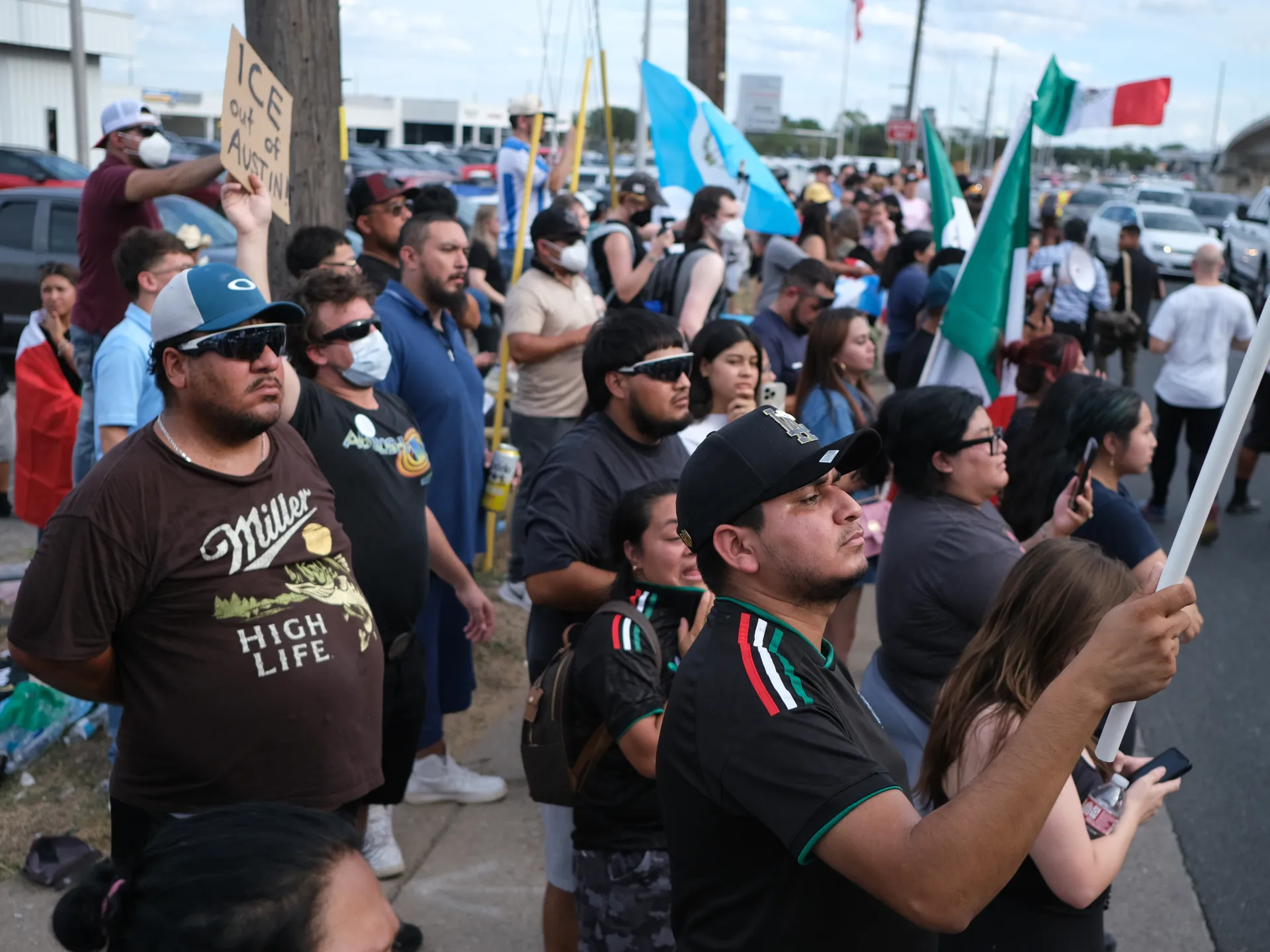 Crowd of demonstrators gathers along a roadside carrying flags and protest signs.