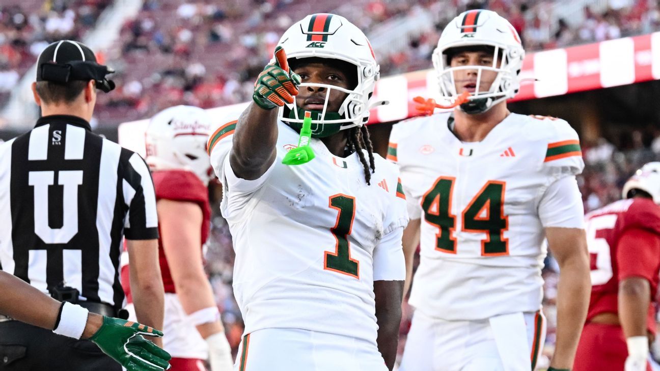 Football player in white uniform points toward the camera as teammates and an official stand nearby