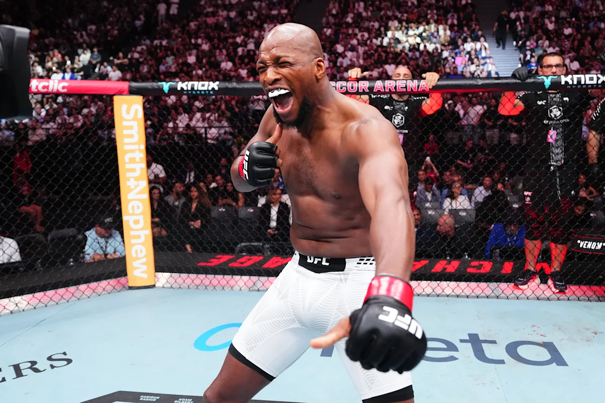 Bald mixed martial artist laughs inside a UFC cage, wearing gloves and white shorts.