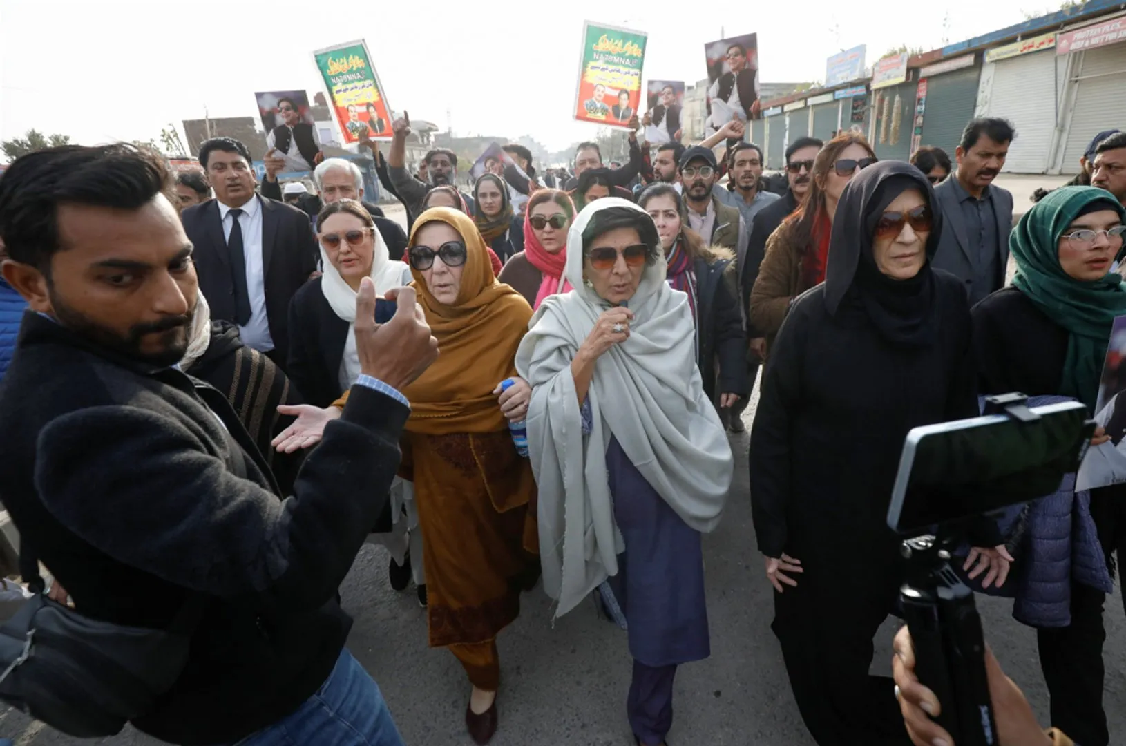 Crowd of protesters, including women in headscarves, march while holding political placards and filming devices.
