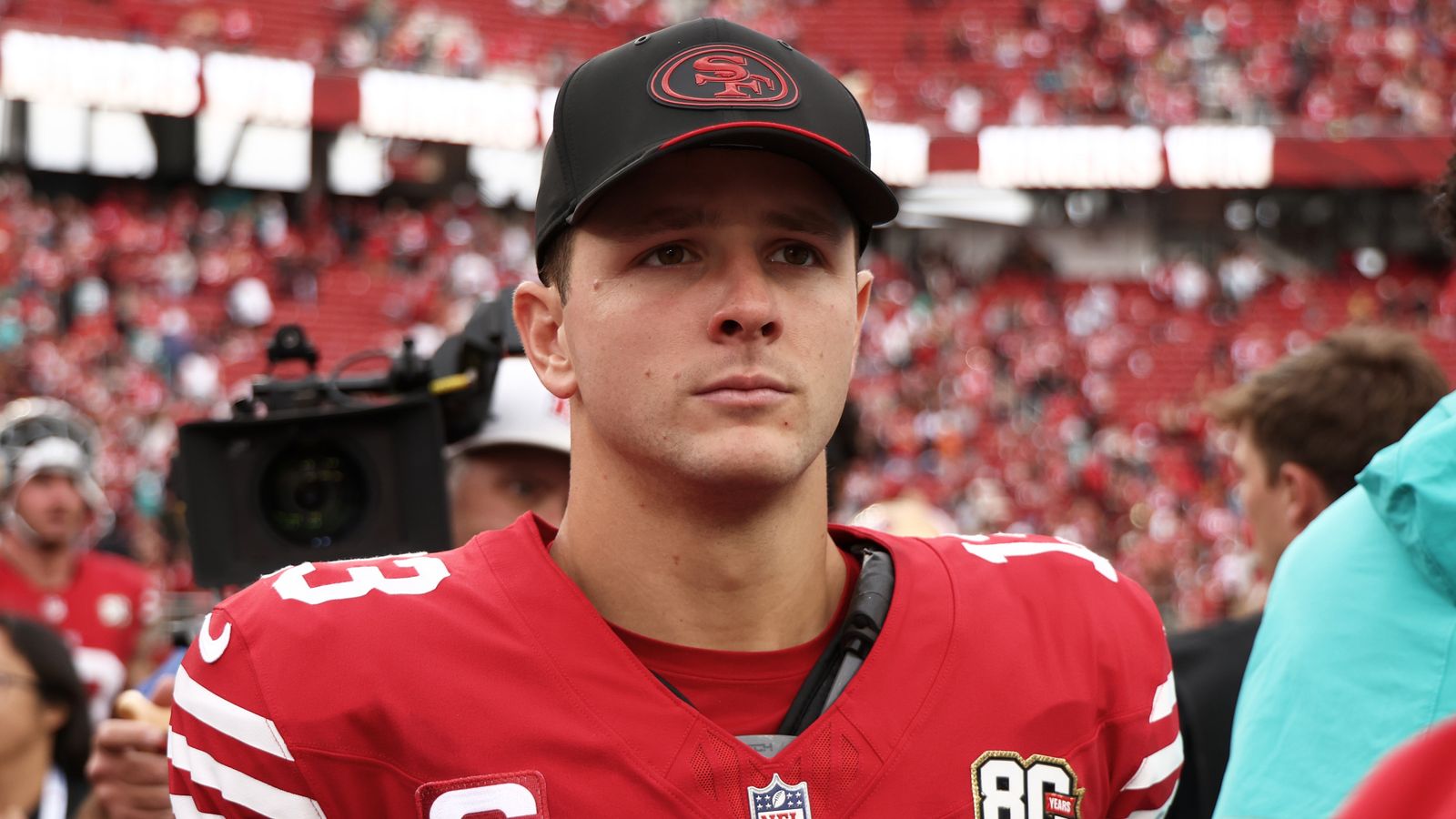 A man in a black cap and red football jersey stands before a blurred crowd.