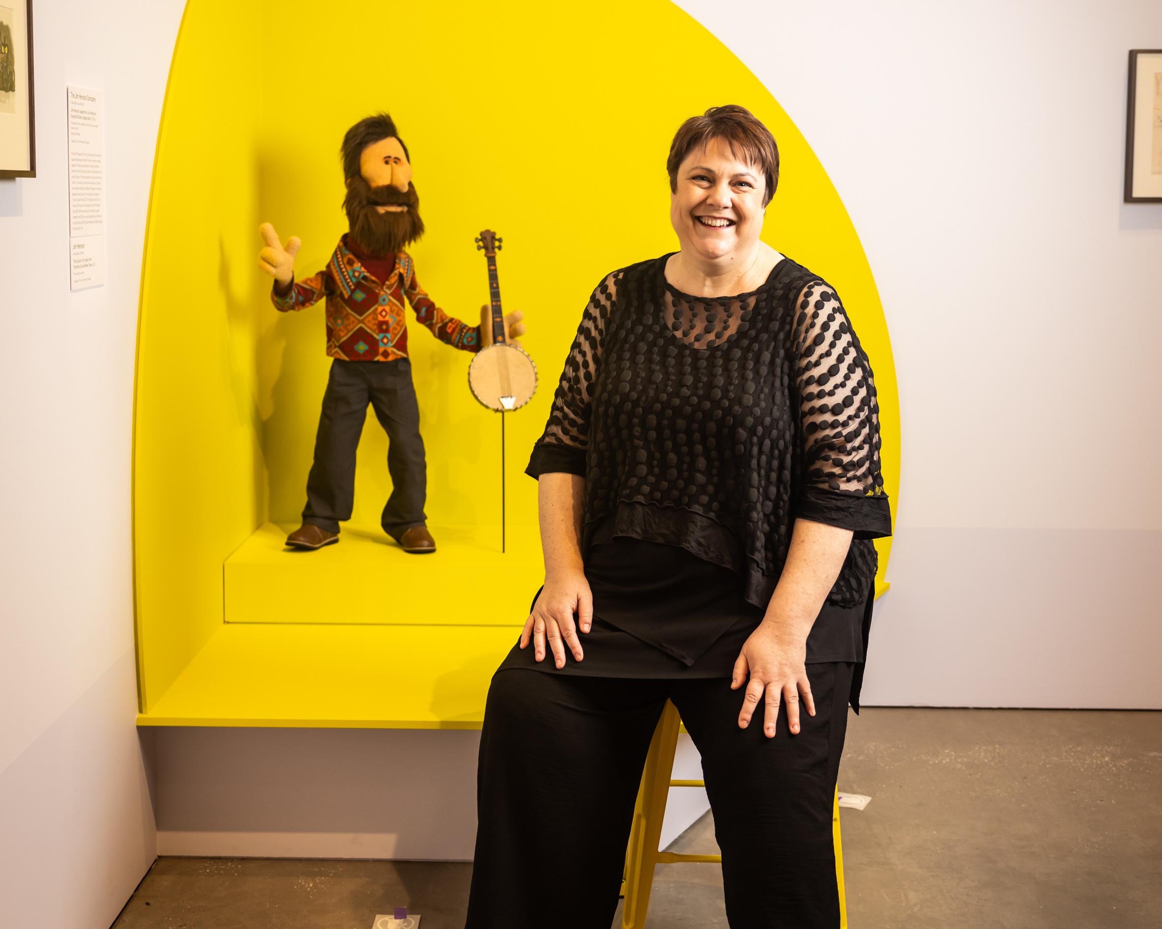 Smiling woman sits beside a bearded puppet holding a banjo in a bright yellow museum display.