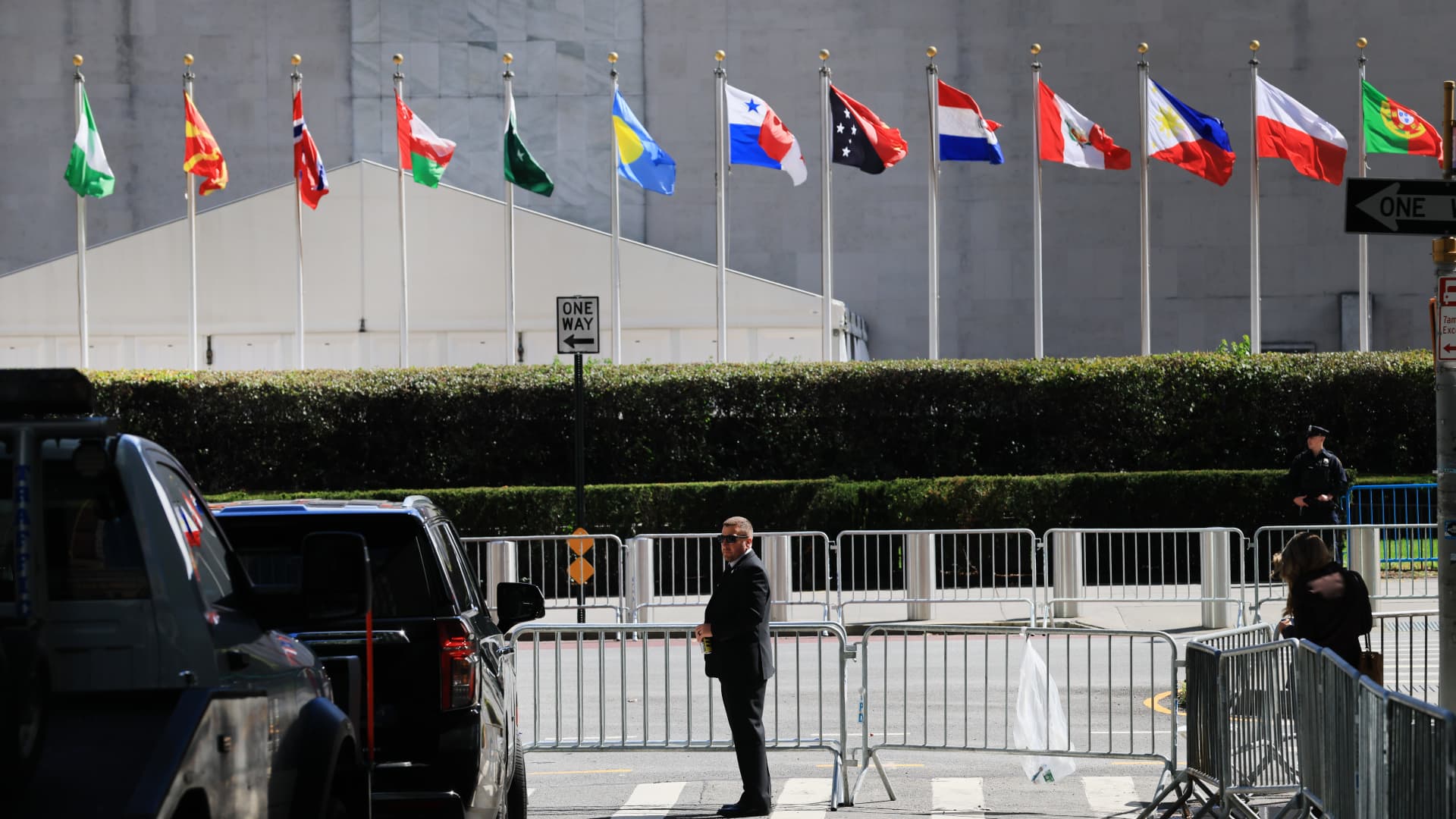 Security barriers and guards line a street beneath a row of national flags outside a large building.