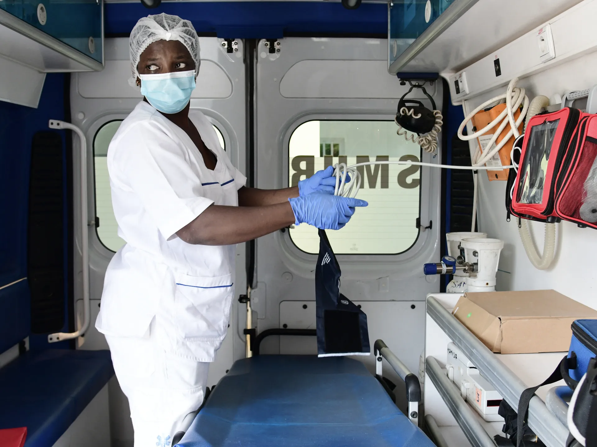 A masked healthcare worker in protective clothing reaches toward medical equipment inside a clinical vehicle.