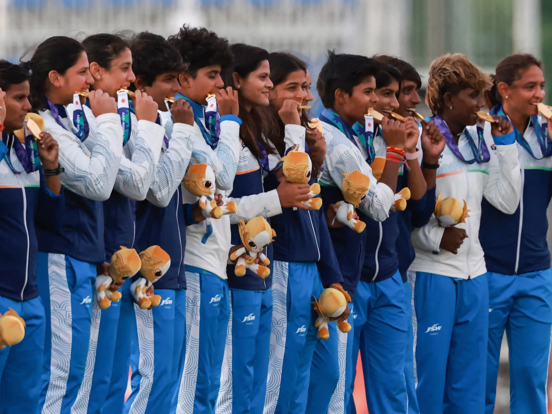 Indian women athletes in blue and white tracksuits bite gold medals while holding mascots.