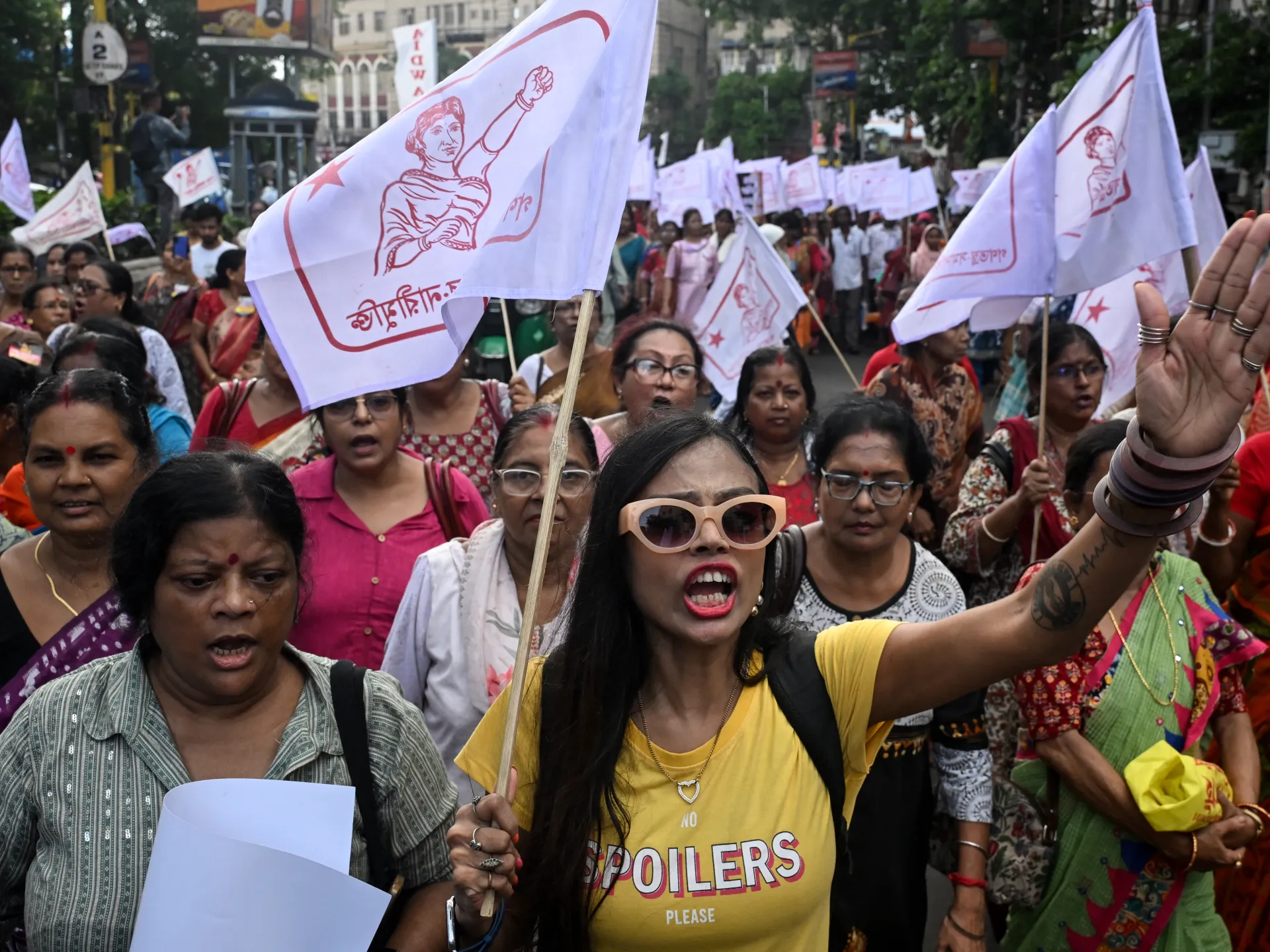 A woman in sunglasses raises her arm during a crowded street demonstration with flags and banners.