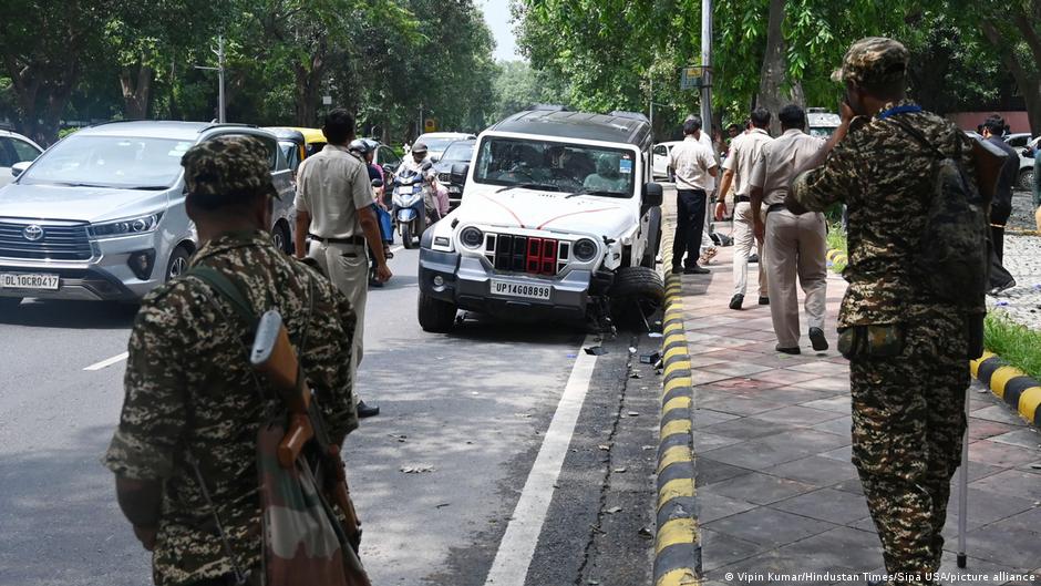 Armed security personnel and police officers surround vehicles along a tree-lined urban roadway.