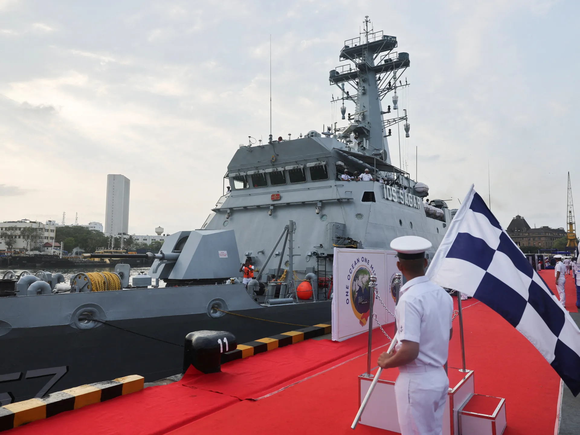 Naval patrol ship docked beside a red carpet as a sailor holds a large blue-and-white flag