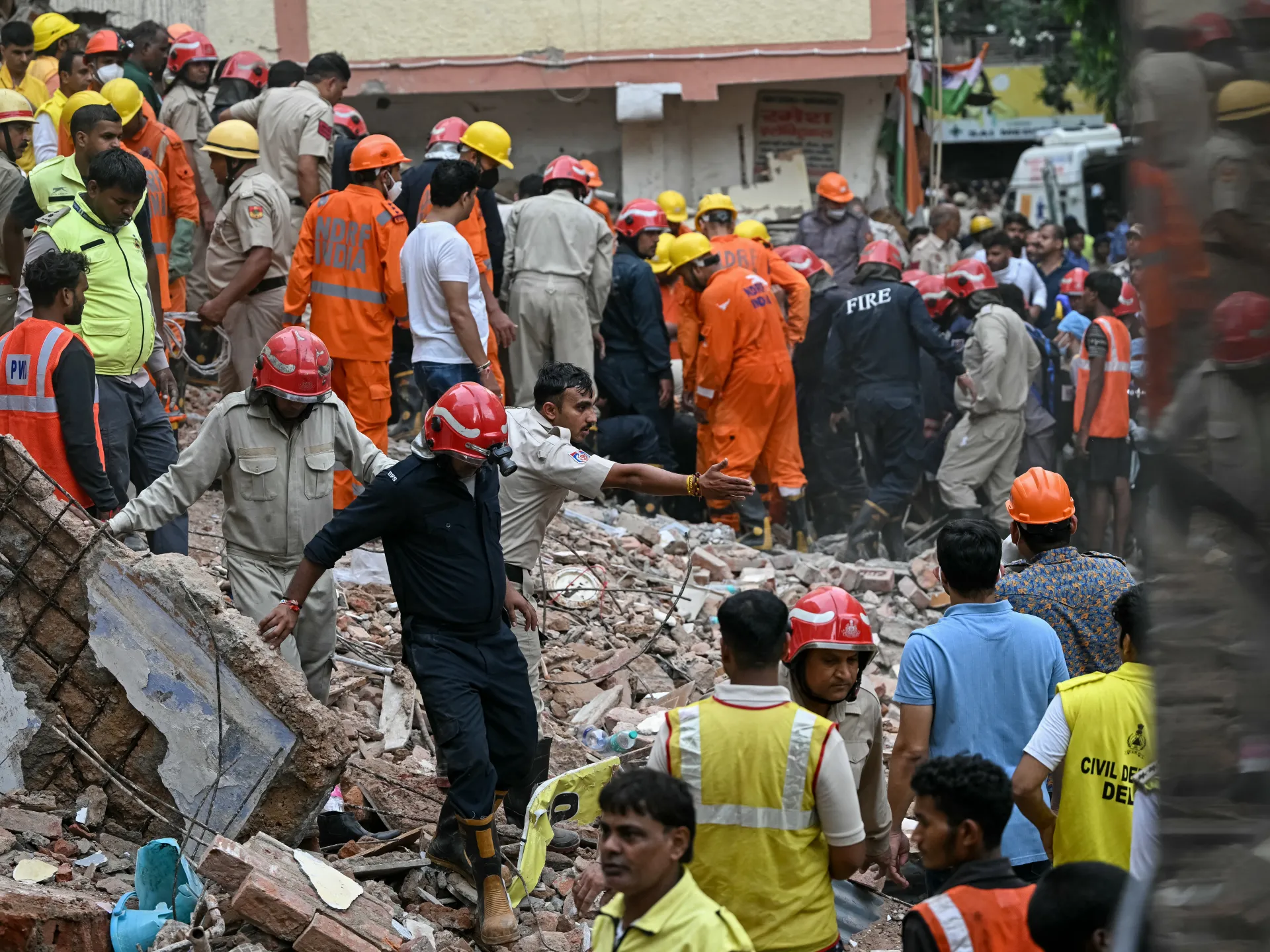 Emergency responders search through rubble as firefighters and civilians gather at a collapsed building site.