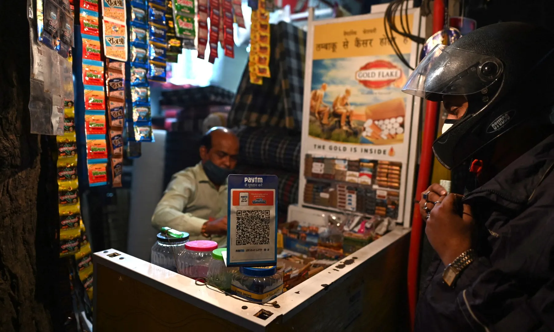 Motorcyclist pays at a small kiosk displaying a Paytm QR code and hanging tobacco packets.