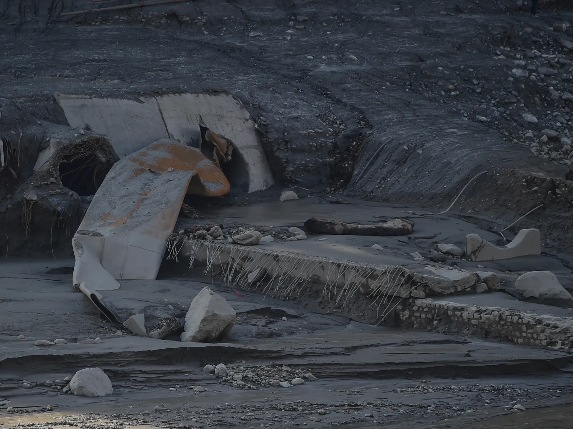 Flood-damaged concrete infrastructure and exposed rebar lie amid dark mud, rocks and debris.