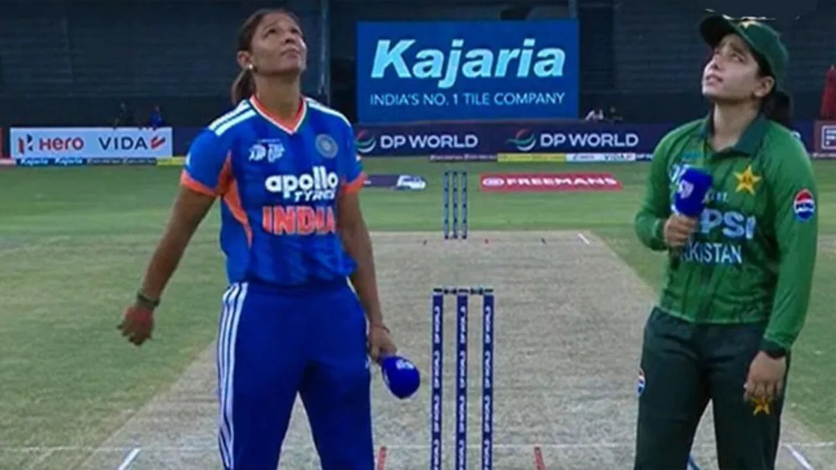 Two women cricket players in India and Pakistan jerseys stand apart during the toss on a cricket pitch.