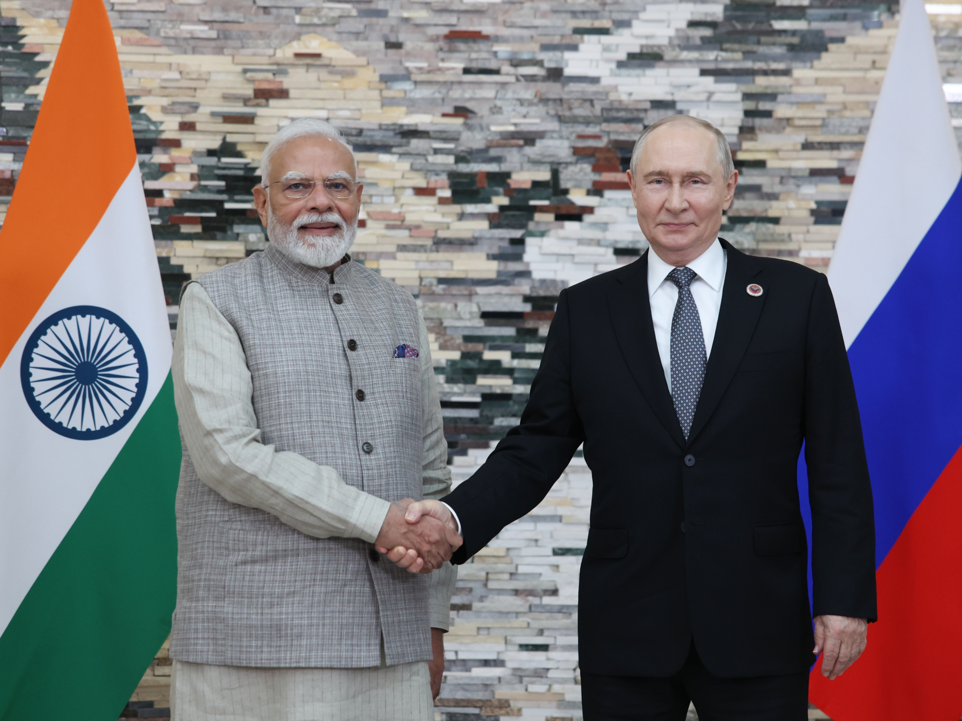 Indian and Russian leaders shake hands between their national flags against a stone wall