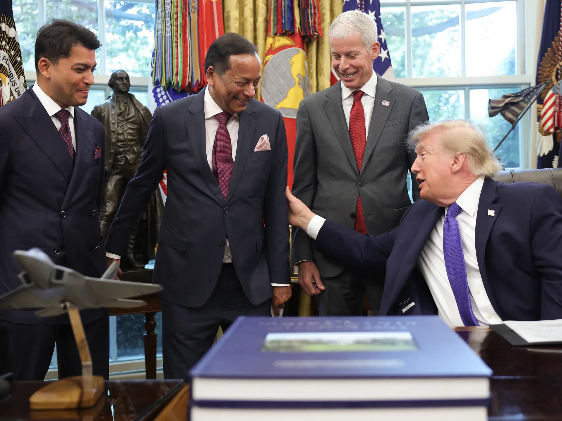 Four suited men gather around a desk as a seated man gestures toward one of them.