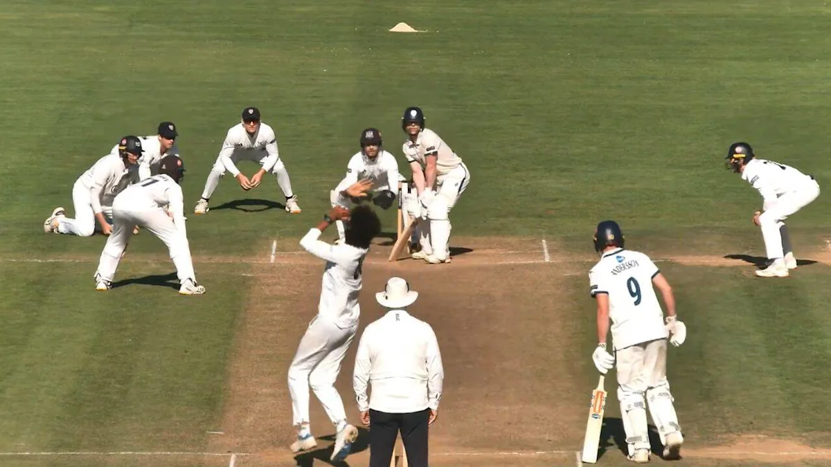 Cricketer delivers a spinning ball as fielders crouch around the batsman on a cricket pitch.