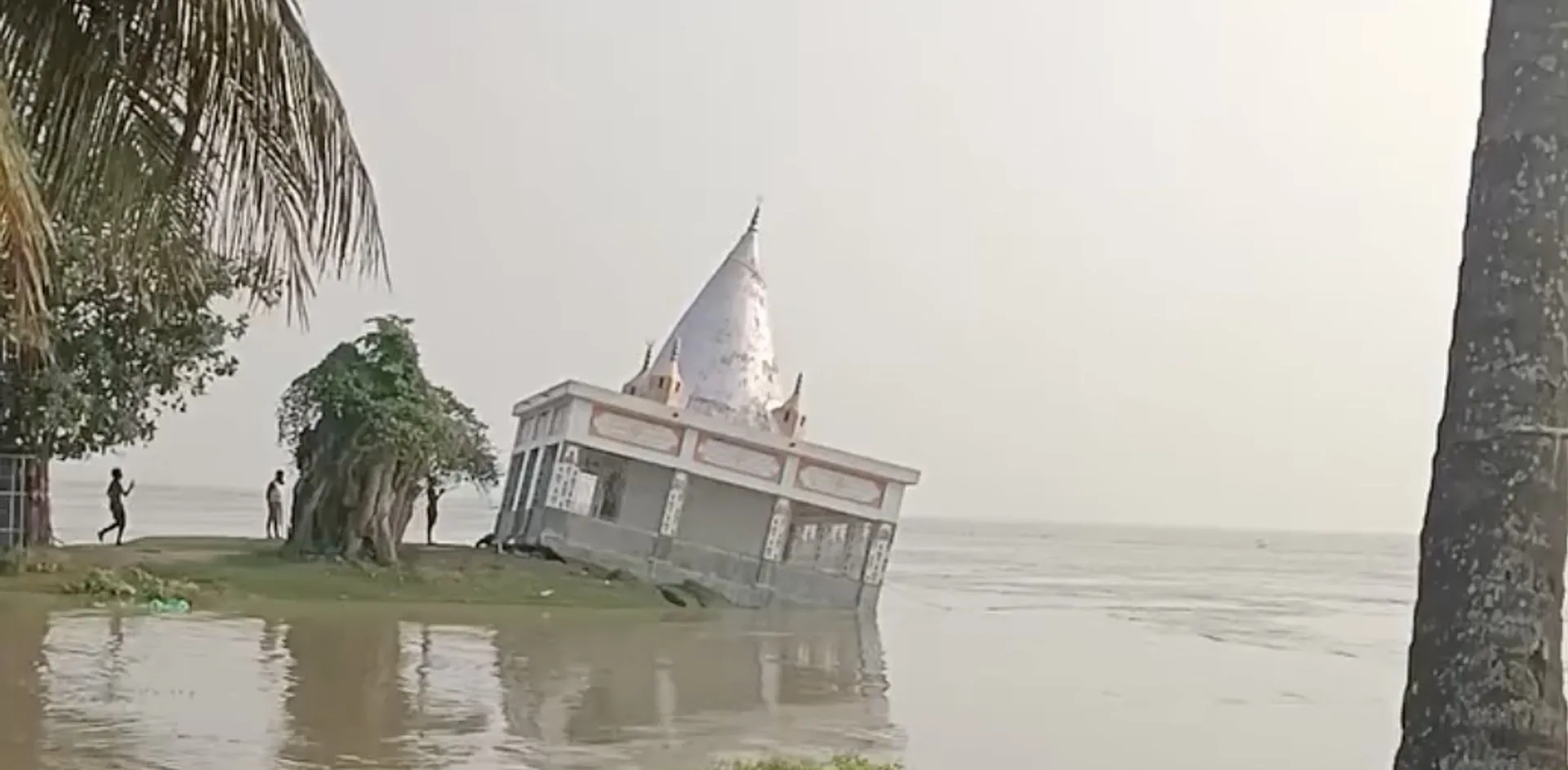 A small white temple stands partially submerged beside a river, with palm trees and people nearby.