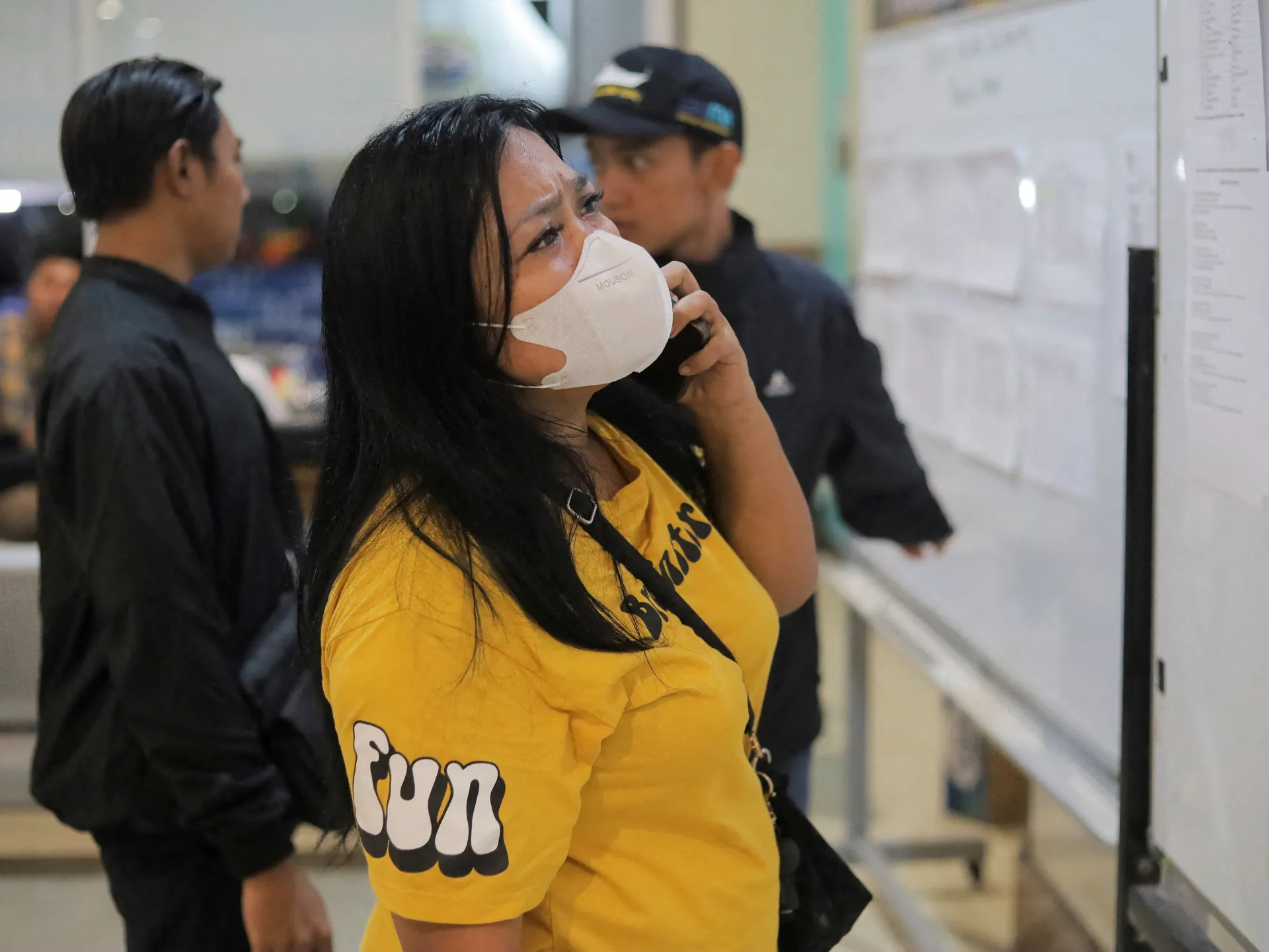 Masked woman in yellow shirt examines a large board as uniformed personnel stand nearby.
