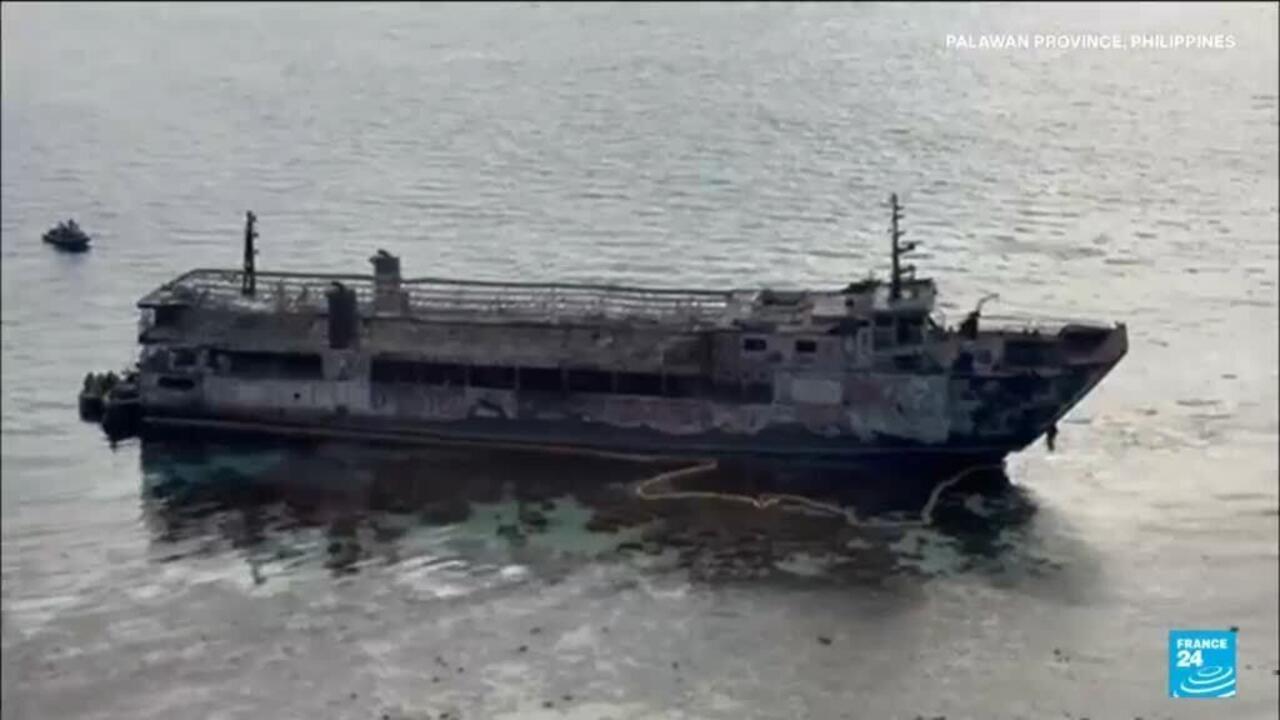 A weathered ferry sits in calm coastal waters, viewed from above, with oil-like dark patches nearby.