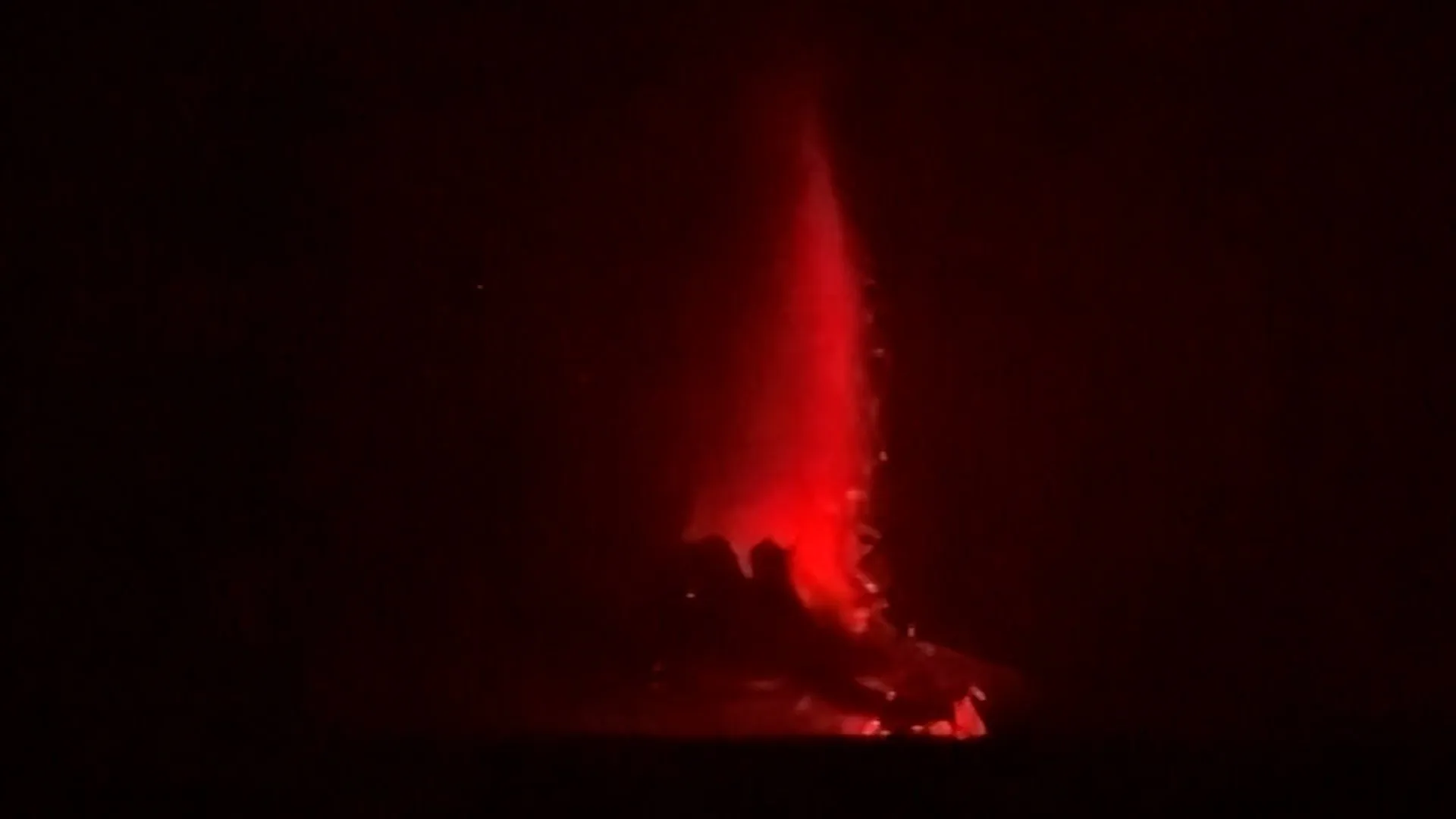 Bright red glow and plume rise from a volcano in darkness at night