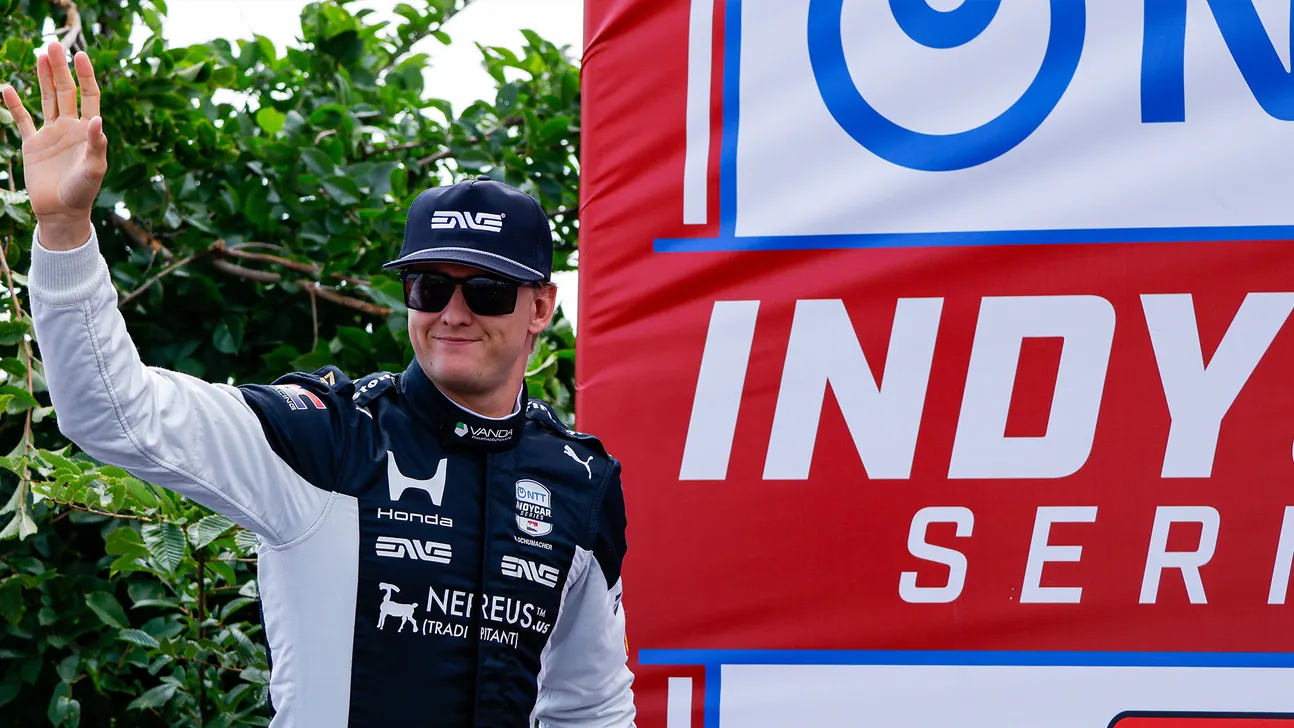 Racing driver in sunglasses and team uniform waves beside a large IndyCar Series sign.