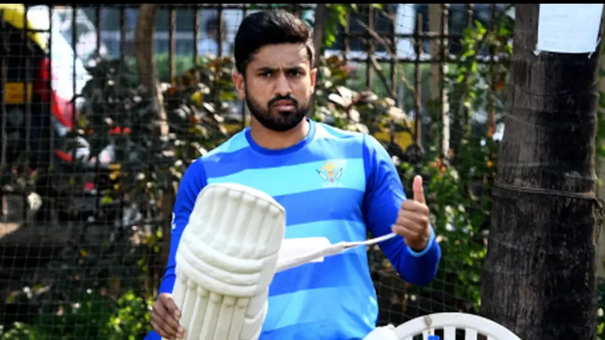 Cricketer in blue jersey holds protective batting pads and gives a thumbs-up outdoors.