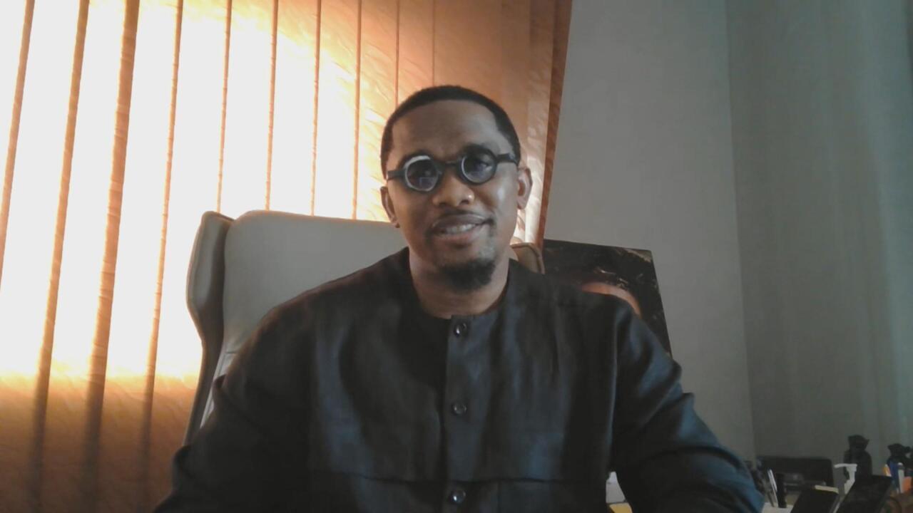 Man wearing round glasses and a dark shirt seated indoors beside a sunlit window