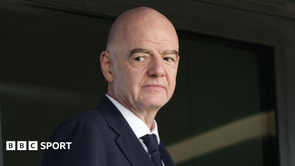 Bald man in a dark suit and tie looking toward the camera indoors