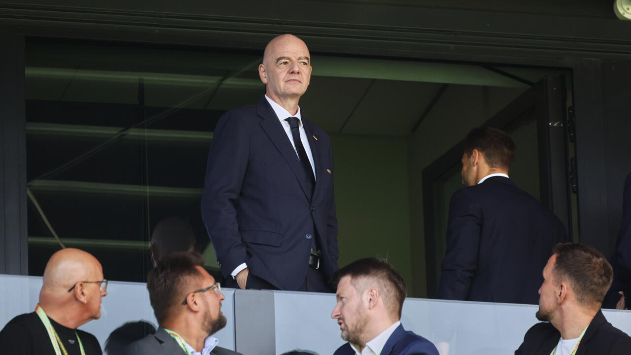 Bald man in dark suit stands in stadium viewing area among seated officials.